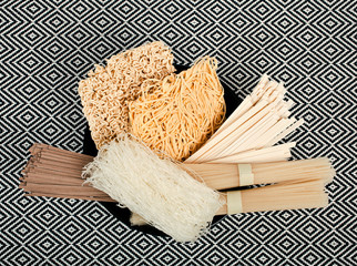 Different bundles of uncooked asian noodles (udon, soba, ramen, rice and glass noodles) in black clay bowl on patterned textile background. Top view.