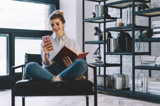 Positive woman resting with book on chair at home stylish interior while reading notification with good news on smartphone, cheerful female blogger watching funny video in blog enjoying free time