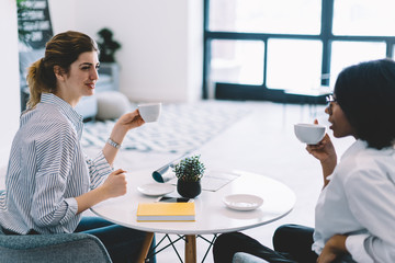 Happy female friends sitting at coffee table and enjoying warm conversations with each other during leisure, positive smiling women taking rest together and gossip during meeting in home interior