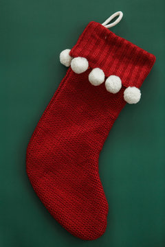 Red knitted Christmas stocking with white pom poms on green background, copy space