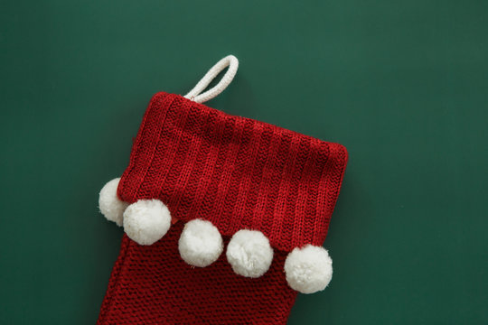 Red knitted Christmas stocking with white pom poms on green background, copy space