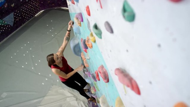 Top shot of fit Caucasian female climber bouldering at vertical indoor climbing wall, grabbing and stepping on hand and foot holds, and padded crash mats on floor below