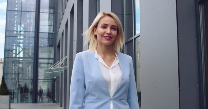 Smiled and suvvessful young blond Caucasian woman in business style walking outdoors at the big urban building and looking at the camera. Portrait shot.