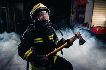 Obraz premium Portrait of a female firefighter while holding an axe and wearing an oxygen mask indoors surrounded by smoke.