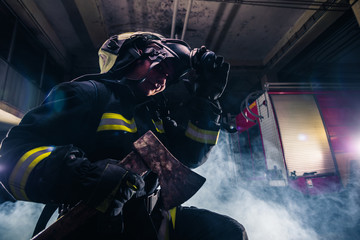 Portrait of a female firefighter while holding an axe and wearing an oxygen mask indoors surrounded by smoke.
