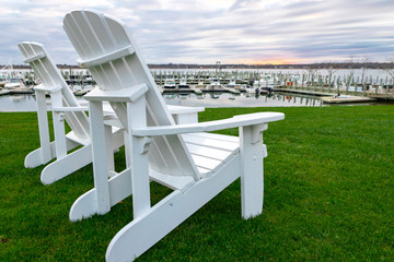 White "Adirondack" style lounge chairs on green grass at waterfront marina, with boats docked in background