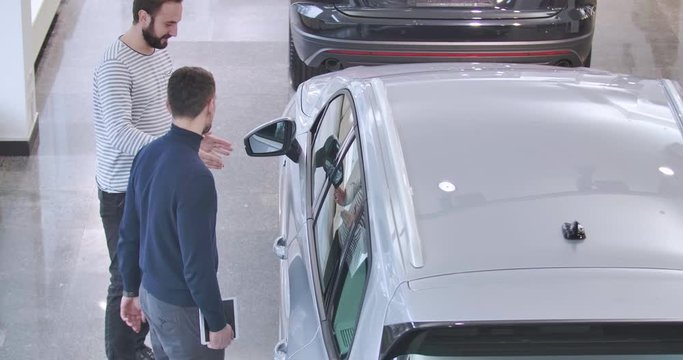 Two Caucasian men standing next to the silver jeep in car dealership and talking. Male trader with tablet and customer discussing advantages of vehicle. Car business. Cinema 4k footage ProRes HQ.