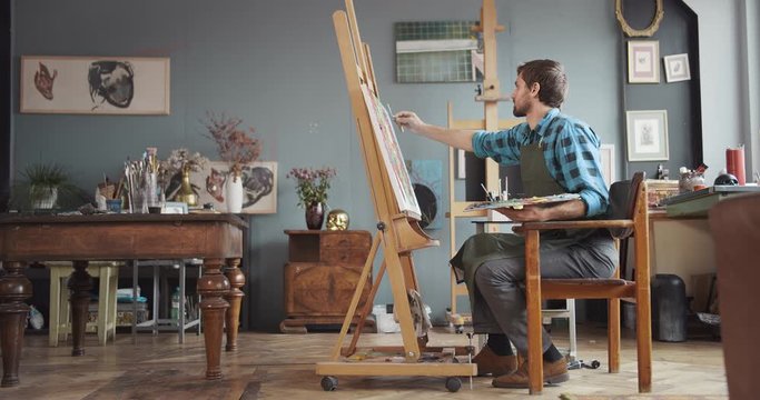 Young male artist working in studio, sitting on old chair near molbert with canvas, lifestyle