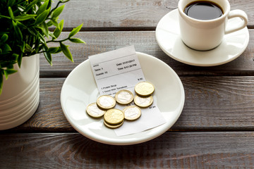 Pay restaurant bill by cash. Reciept and coins on plate on dark wooden background