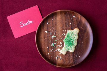 Christmas note to Santa, sugar cookie, half eaten, and crumbs on a wood plate, red background
