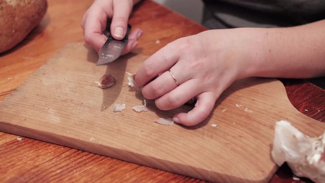 Woman's hands with a knife crush the garlic on a chopping board, then peel the skin. Preparations for making a sandwich with garlic. Close up.