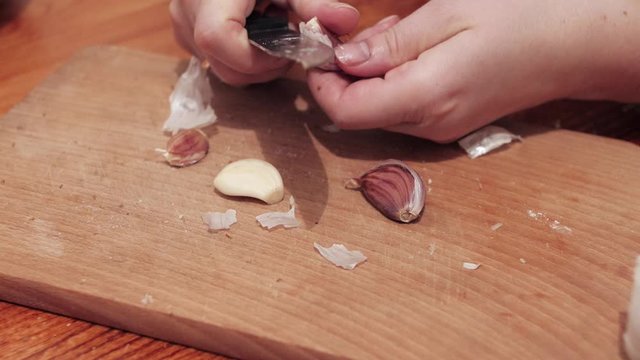 Woman's hands with a knife crush the last clove of garlic on a chopping board, then peel the skin. Moves skins off the board. Preparations for making a sandwich with garlic.