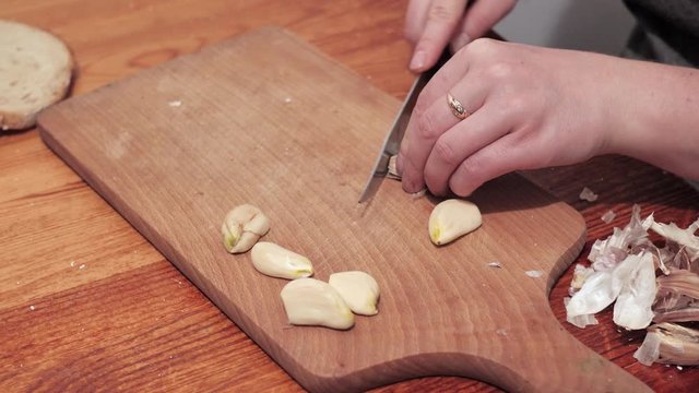 Woman's hands with a knife crush the last clove of garlic on a chopping board, then peel the skin. Moves skins off the board. Preparations for making a sandwich with garlic.