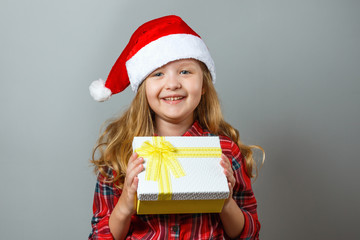 Christmas and New Year concept. Cute cheerful child holds a box with a gift in his hand. Charming little girl in santa hat and red dress on a gray background