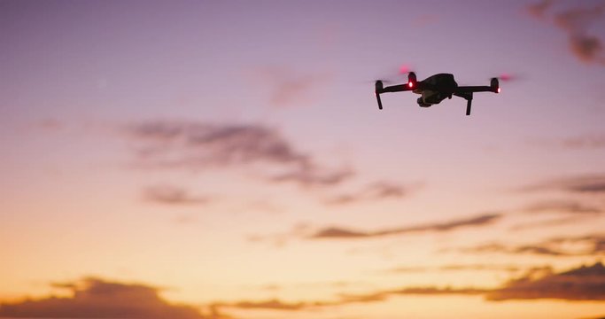 Orbiting view of a quadcopter drone silhouette hovering and flying at sunset with a purple yellow sky