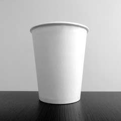 White paper disposable glass on  table on background of white stanf. Square shot. Black and white photo. Front view.