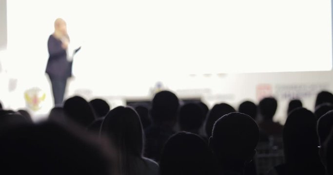 Unrecognizable businesswoman on stage talking microphone ans showing presentation on screen to large audience sitting back to camera, blur background. Training, conference, public speech concept.