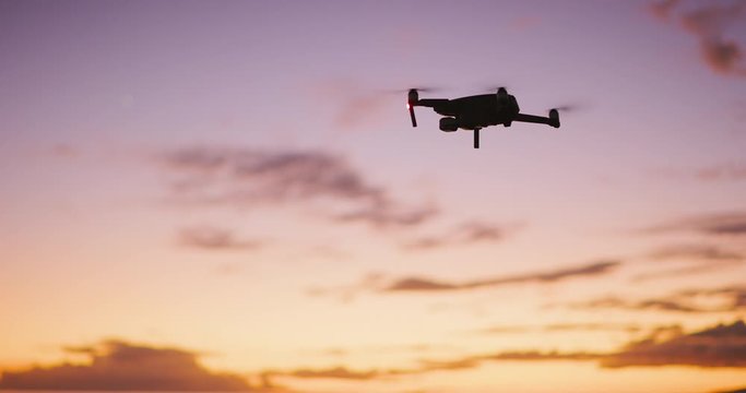 Orbiting view of a quadcopter drone silhouette hovering and flying at sunset with a purple yellow sky