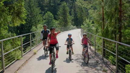 Family of Caucasian parents and children in bicycles crossing a bridge together in Slovenia, Drone flies up at the end to let them pass - Powered by Adobe