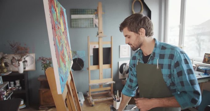 Handsome man looking at picture and rubbing hands, artist satisfied with painting, inspiration