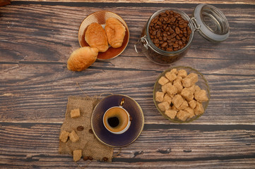 A Cup of coffee, pieces of brown sugar in a sugar bowl, coffee beans in a glass jar and croissants on a wooden background.