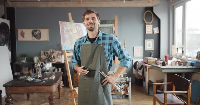 Young handsome male painter looking at finished work, turning to camera and smiling, holding brushes