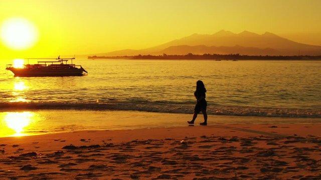 Woman Walking Under The Beautiful Setting Sun On The Beach Of Bora Bora With Mountains And Tourist Boat In The Background - Wide Shot