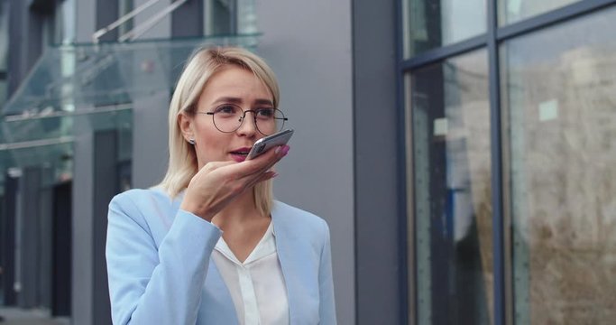 Caucasian young beautiful busy businesswoman walking outdoors in the morning and recording a voice message while talking to the phone speaker. Close up.