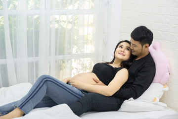A beautiful pregnant woman and her handsome husband are smiling while spending time together in bed. A man is listening to the sound of a baby in his stomach.