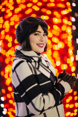 Smiling brunette in checkered coat and black hat on red background with glowing yellow lights