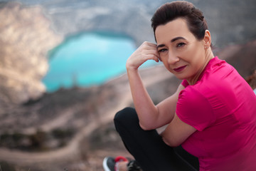 Fototapeta na wymiar Portrait of 40s years traveler woman dressed sportswear, pink topic and black fitness leggings, sit on the top of mountain against the heart shaped quarry.