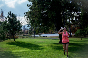 40s years old active woman dressed sportwear and  headphones  is running and drink water outdoor in the park with green grass. Mature woman living healthy lifestyle