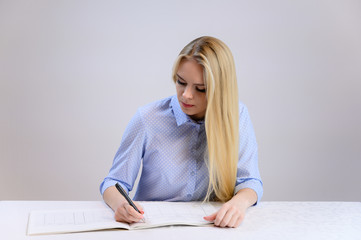 Concept cute model student secretary works sitting at a table. Close-up portrait of a beautiful blonde girl with excellent makeup with long smooth hair on a white background in a blue shirt.