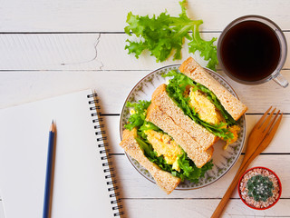Scrambled eggs sandwich breakfast, a cup of black coffee and empty notebook paper top view on white wooden table.