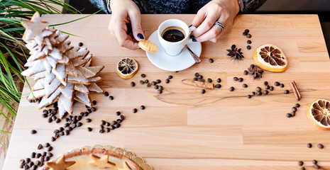 Hands of woman holding cup of coffee with biscuit on a wooden background with copy space. Christmas decoration with sweets in baker. Merry Christmas. Panoramic banner.