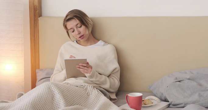Thoughtful young woman writing notes sitting in bed at home