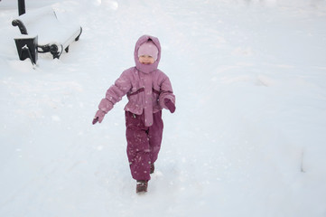 Fototapeta na wymiar Winter in the park. A toddler girl wearing warm clothes walking through snowfalll in the park