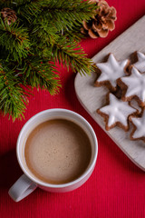 Obraz na płótnie Canvas Cup of coffee on a red textile background with twig of decorative christmas tree and christmas cookies (cinnamon stars, Zimtsterne in German). Selective focus, top view, close-up. Christmas wallpaper.