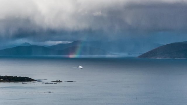 A ferry crossing from Bognes to Lodingen. The fjord is calm, thick white-grey clouds hanging above, a rainbow appears in a distance. High mountains and green fields are framing the scene.