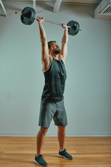 Portrait of super fit muscular young man working out in gym with barbell on gray background,...