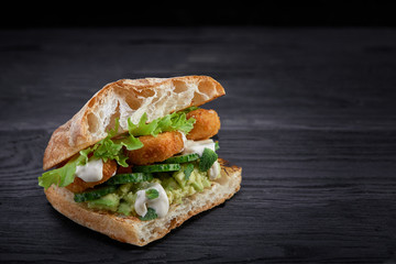 Appetizing sandwich on a wooden board. Baguette sandwich with filling from lettuce, slices tomato. Dark wooden background. View from above. Close-up. Macro photography.