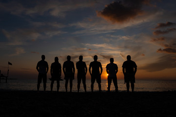 Silhouettes of men standing by the sea against the sunset. Group of unrecognizable men with...