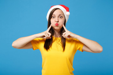 Funny young woman Santa girl in yellow t-shirt Christmas hat posing isolated on blue background. New Year 2020 celebration holiday concept. Mock up copy space. Point index fingers on blowing cheeks.