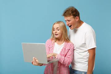 Young amazed couple friends guy girl in white pink empty blank design t-shirts posing isolated on pastel blue background. People lifestyle concept. Mock up copy space. Working on laptop pc computer.