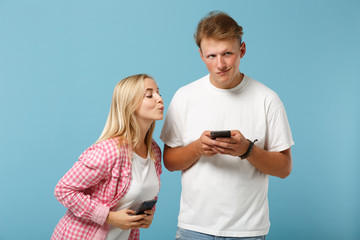 Young curious couple friends guy girl in white pink empty blank t-shirts posing isolated on pastel blue background. People lifestyle concept. Mock up copy space. Using mobile phone typing sms message.