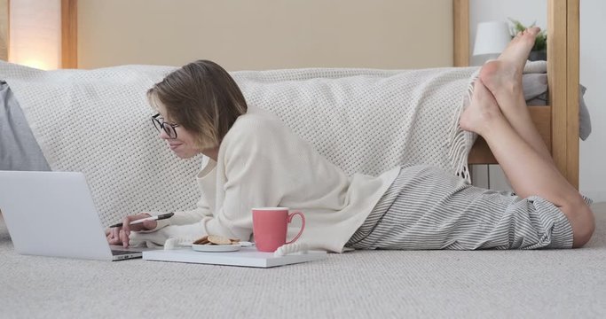 Young woman in pajamas having breakfast while working on laptop lying on carpet in bedroom