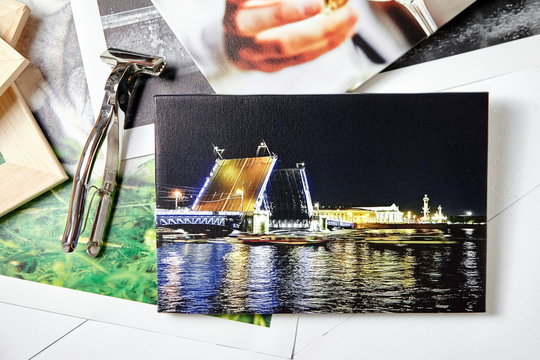 Canvas prints, colorful photos of St. Petersburg and drinking people, tool for wrapping. Photographs, stretcher bars and canvas pliers on white wooden table. Photos printed on glossy synthetic canvas
