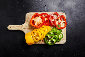 Cutting board with slices of red, green, yellow bell peppers on black background. Sliced sweet peppers in different colors on stone table, vegetable salad ingredient, cooking healthy food, top view