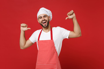 Cheerful young bearded male chef cook or baker man in striped apron white t-shirt toque chefs hat posing isolated on red background. Cooking food concept. Mock up copy space. Point thumbs on himself.