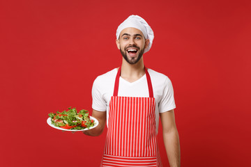 Excited young bearded male chef cook or baker man in striped apron toque chefs hat posing isolated on red wall background. Cooking food concept. Mock up copy space. Hold plate with vegetable salad.